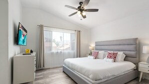 Une chambre d’hôtel moderne avec un grand lit, un ventilateur de plafond, un téléviseur posé sur une commode blanche et une vue sur l’extérieur par une grande fenêtre.