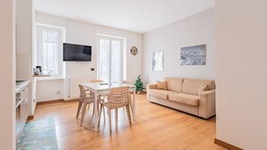 Una sala de estar moderna con un sofá color beige, una mesa de comedor con sillas de mimbre, un televisor de pantalla plana en la pared y un pequeño árbol de Navidad.