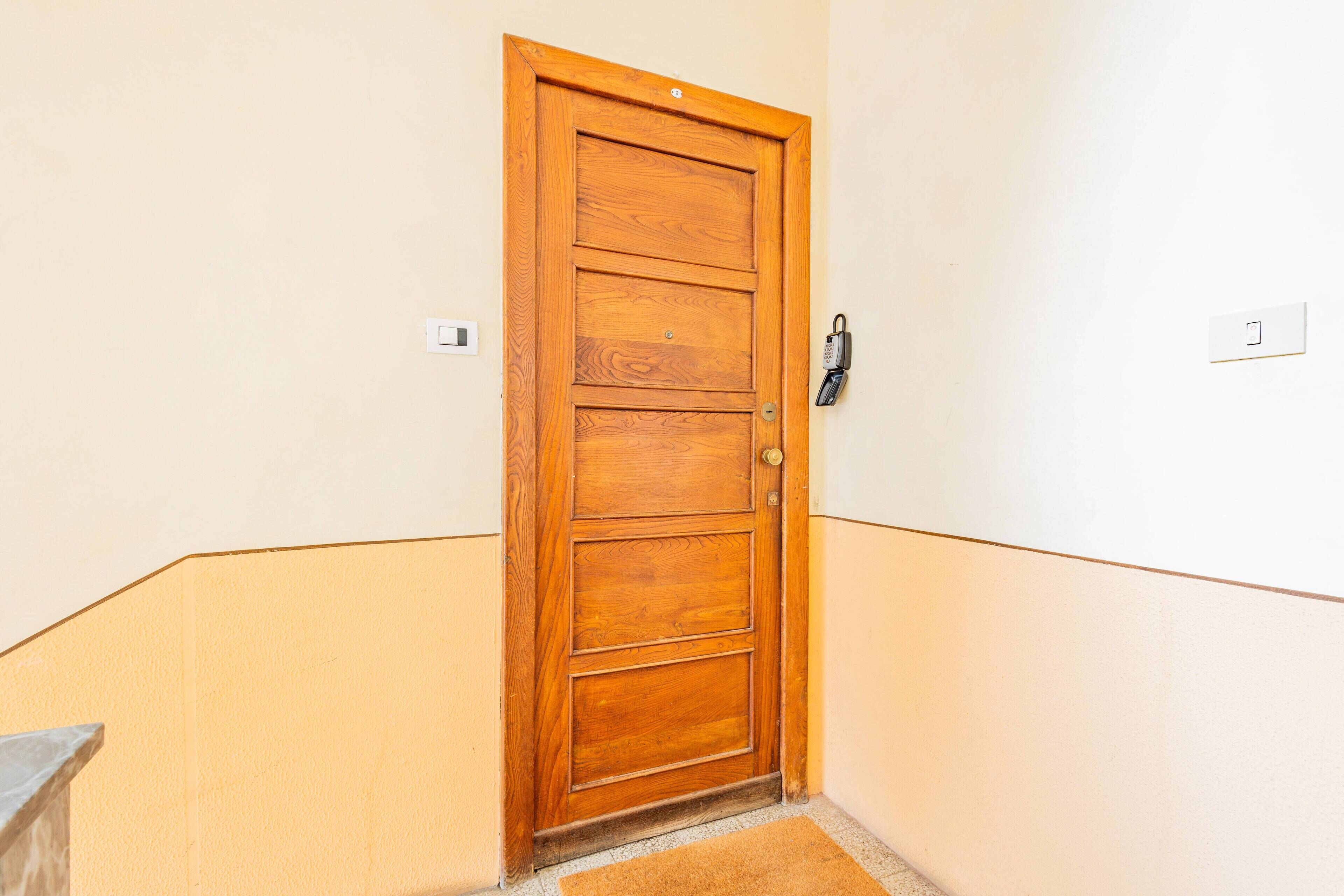 Una porta di legno con serratura a keypad, un tappetino d'ingresso e un interruttore sulla parete.
