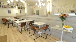 A dining area with white tables and brown chairs, a large mirror, and a vase with flowers on a small table.