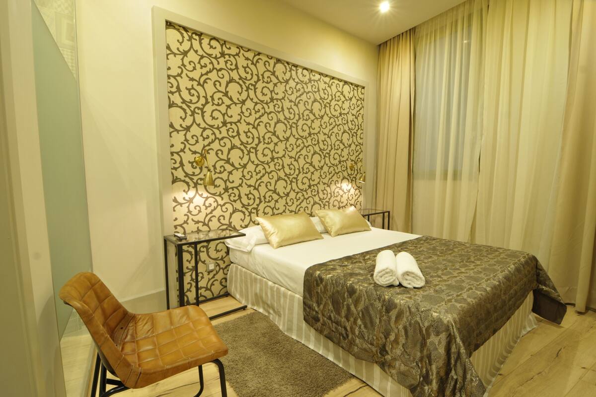 A hotel room with a bed, a chair, a nightstand, and a patterned wall.