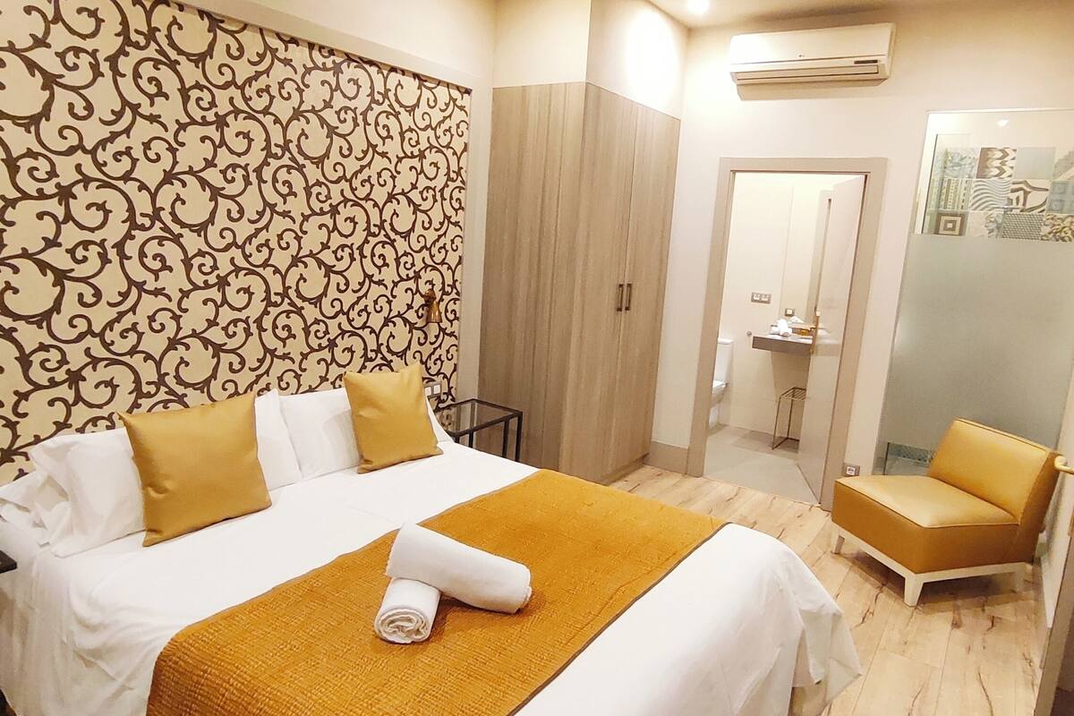 A hotel room with a patterned wallpaper, a bed with white and yellow bedding, a small table with a glass top, a chair, and a television.