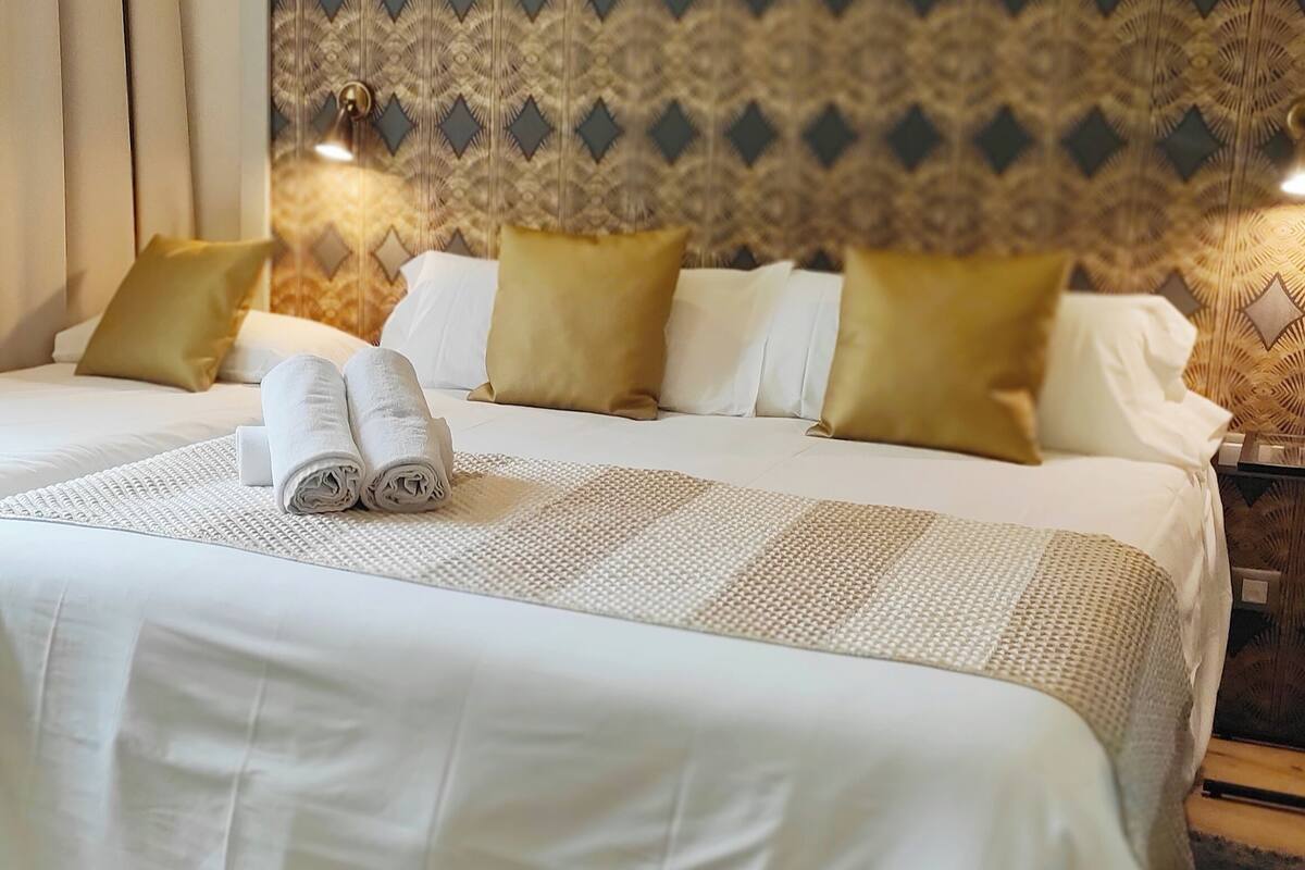 A neatly made bed with white linens and rolled towels, a patterned headboard, and a bedside table with a lamp.