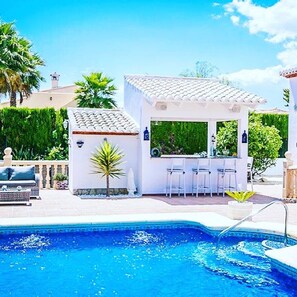 A pool area with a bar and seating, surrounded by greenery and a clear sky.