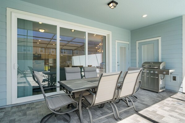 An outdoor patio with a table and chairs, a grill, and a sliding glass door leading to a covered area. - Cozy 5BR Home With Pool Fun Themed Rooms (Clermont)