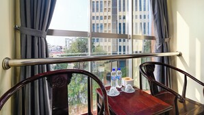 Ein Zimmer mit Blick auf ein hohes Gebäude, ein Fenster mit Vorhängen, ein Holzstuhl und ein Tisch mit Wasserflaschen und einer Taschentuchbox.