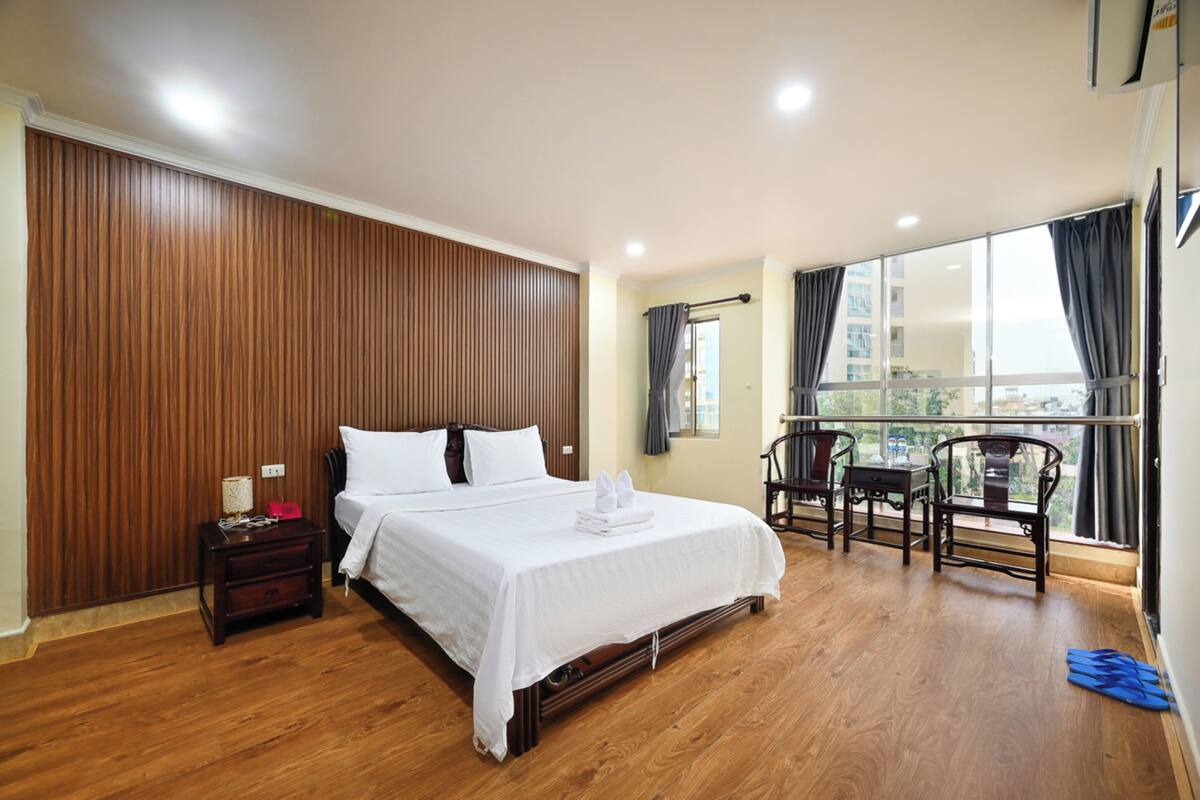 Ein Hotelzimmer mit einer Wandpaneele aus Holz, einem großen Bett mit weißer Bettwäsche, einem kleinen Tisch mit Stühlen und Blick durch ein großes Fenster.
