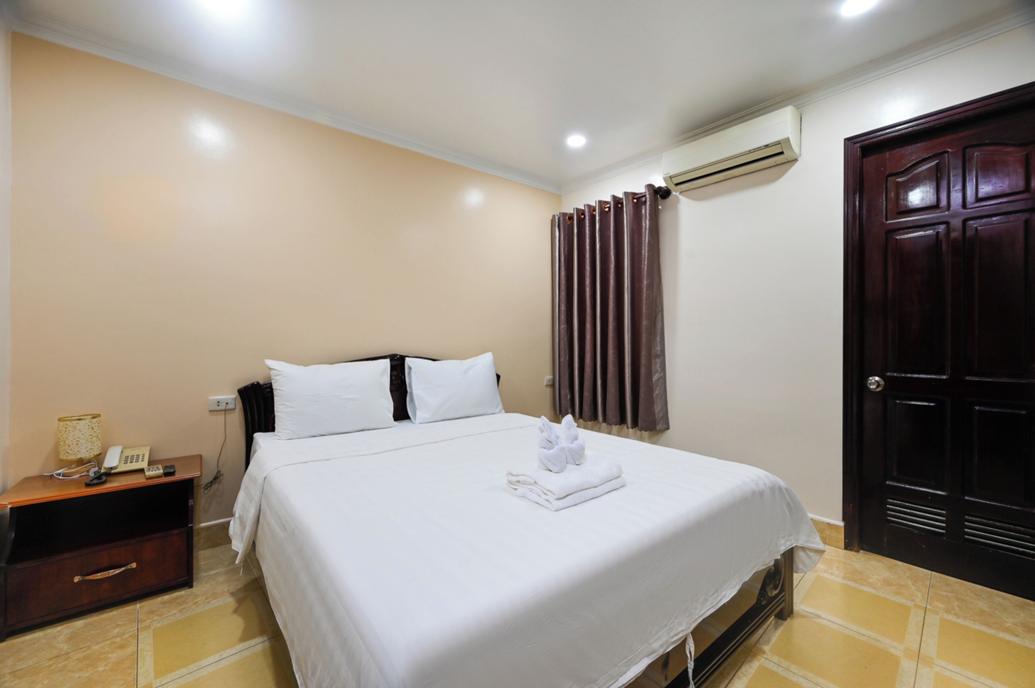 Una habitación de hotel con una cama, una mesita de noche, un equipo de aire acondicionado y una puerta de madera oscura.