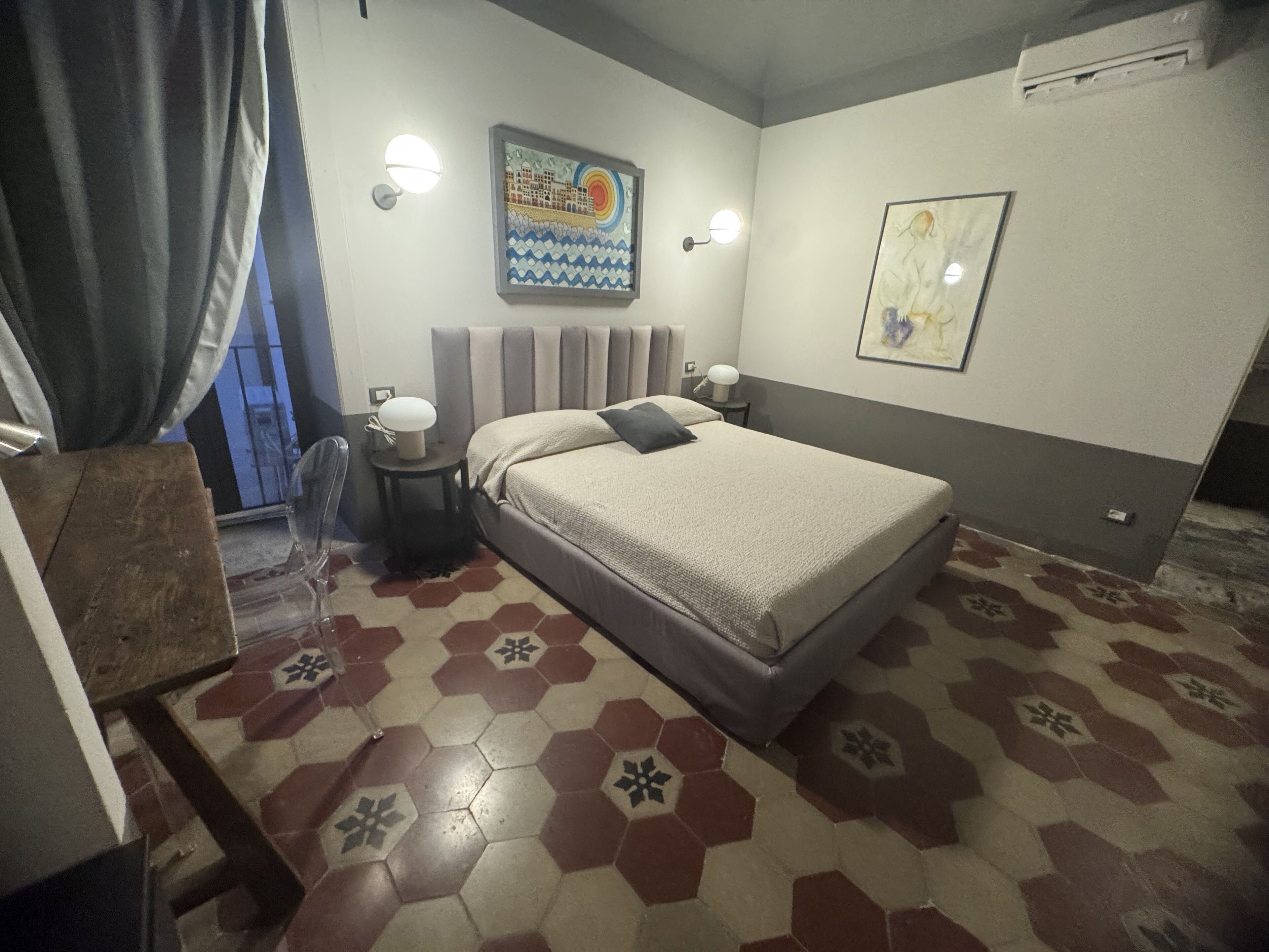Quarto com cama, duas mesas de cabeceira, luminária na parede, um quadro e piso estampado.