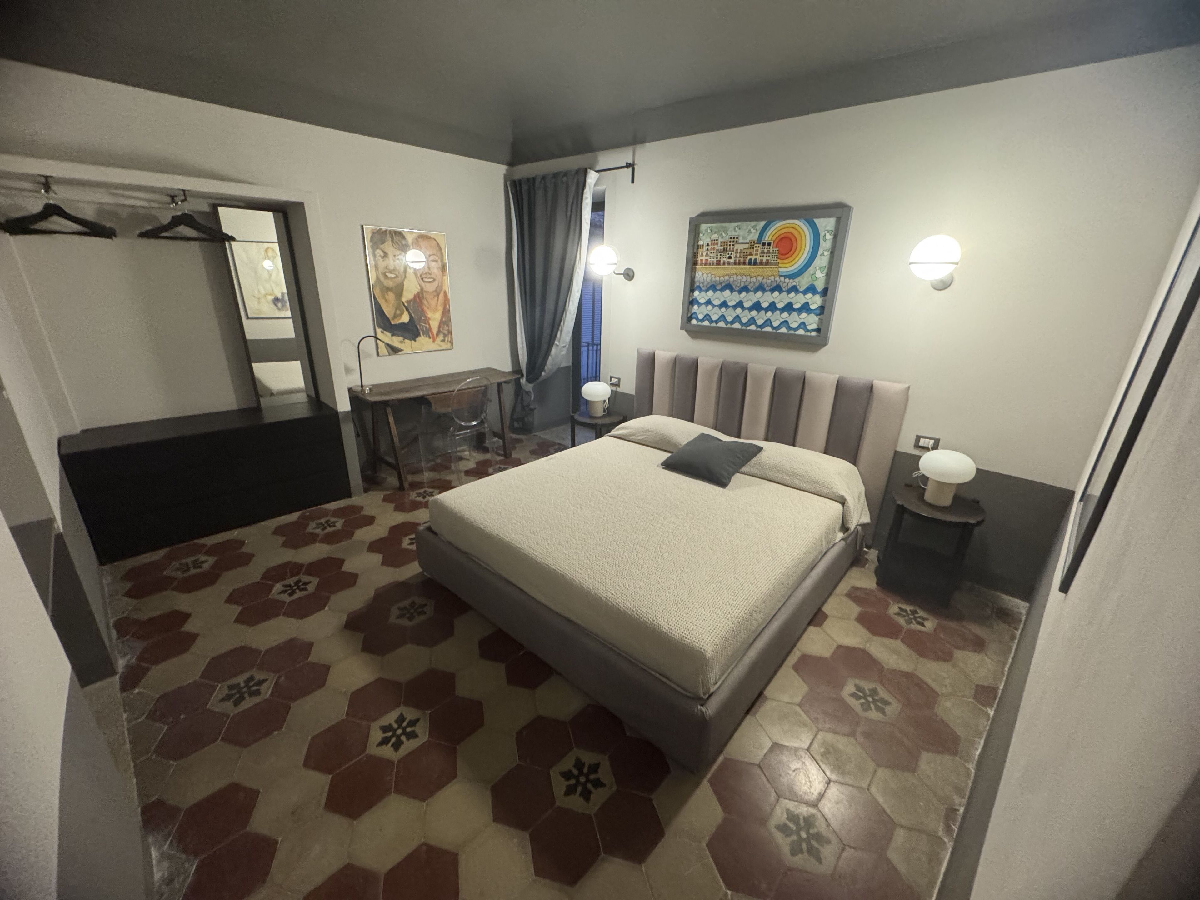 A hotel room with a bed, a desk, a chair, a wardrobe, a mirror, a painting, and a nightstand with a lamp.