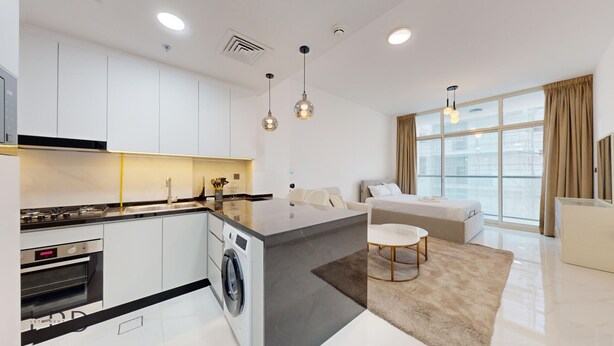 A modern kitchen with a central island, built-in oven, and washing machine. There is a dining area with a round table and chairs. The room has large windows with curtains and a view of the city. - Modern Furnished Studio in Uniestate Supreme (Dubai)