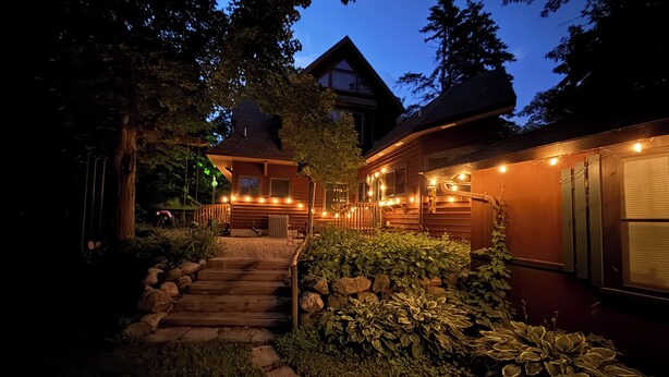 A wooden house with a stone staircase leading to a front porch, surrounded by greenery and string lights.