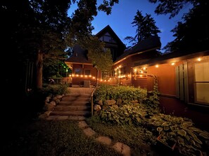 A wooden house with a stone staircase leading to a front porch, surrounded by greenery and string lights.