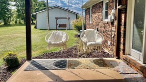 Een overdekt terras met twee witte wicker stoelen en een schommel, een gevel van baksteen met een 'WELCOME' bord, en een heldere lucht.