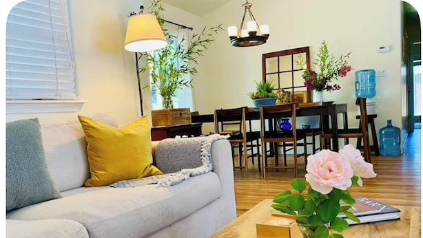Una sala de estar con un sofá, una mesa de centro con un florero, un área de comedor con una mesa y sillas, y un ventanal con persianas.