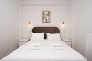 A neatly made bed with white linens and two pillows, a brown headboard, bedside tables with lamps, and a framed picture on the wall. - LIV - Captivating Apartment in Pagrati (Athens)