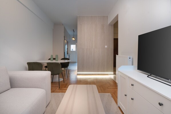 A modern living room with a sofa, a flat-screen TV, a dining area with chairs, and a kitchen in the background. - LIV - Captivating Apartment in Pagrati (Athens)