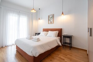 A hotel room with a large bed, two bedside tables, a nightstand, a framed picture on the wall, and a window with sheer curtains. - LIV - Captivating Apartment in Pagrati (Athens)