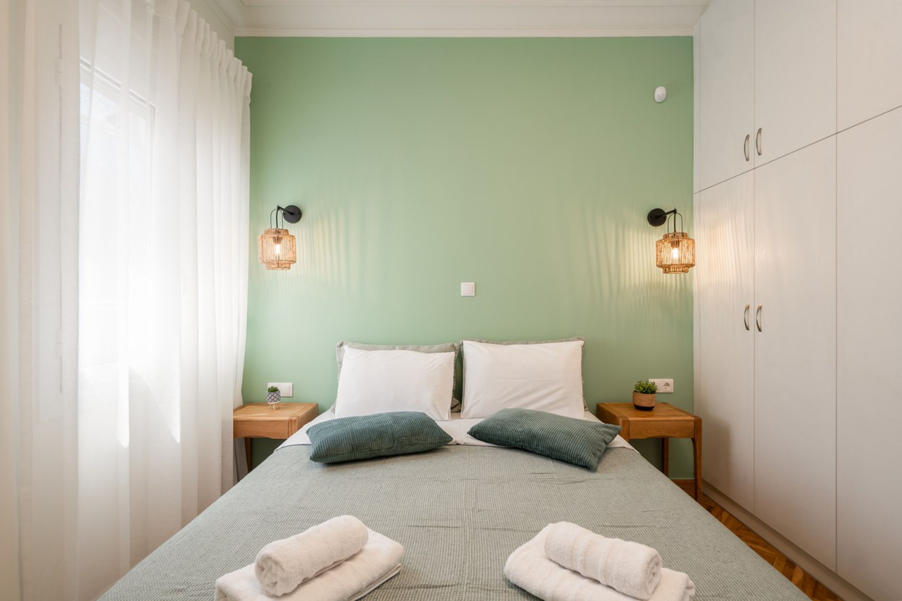A neatly made bed with pillows and towels, a bedside table with a plant, and a wardrobe in a room with light green walls.