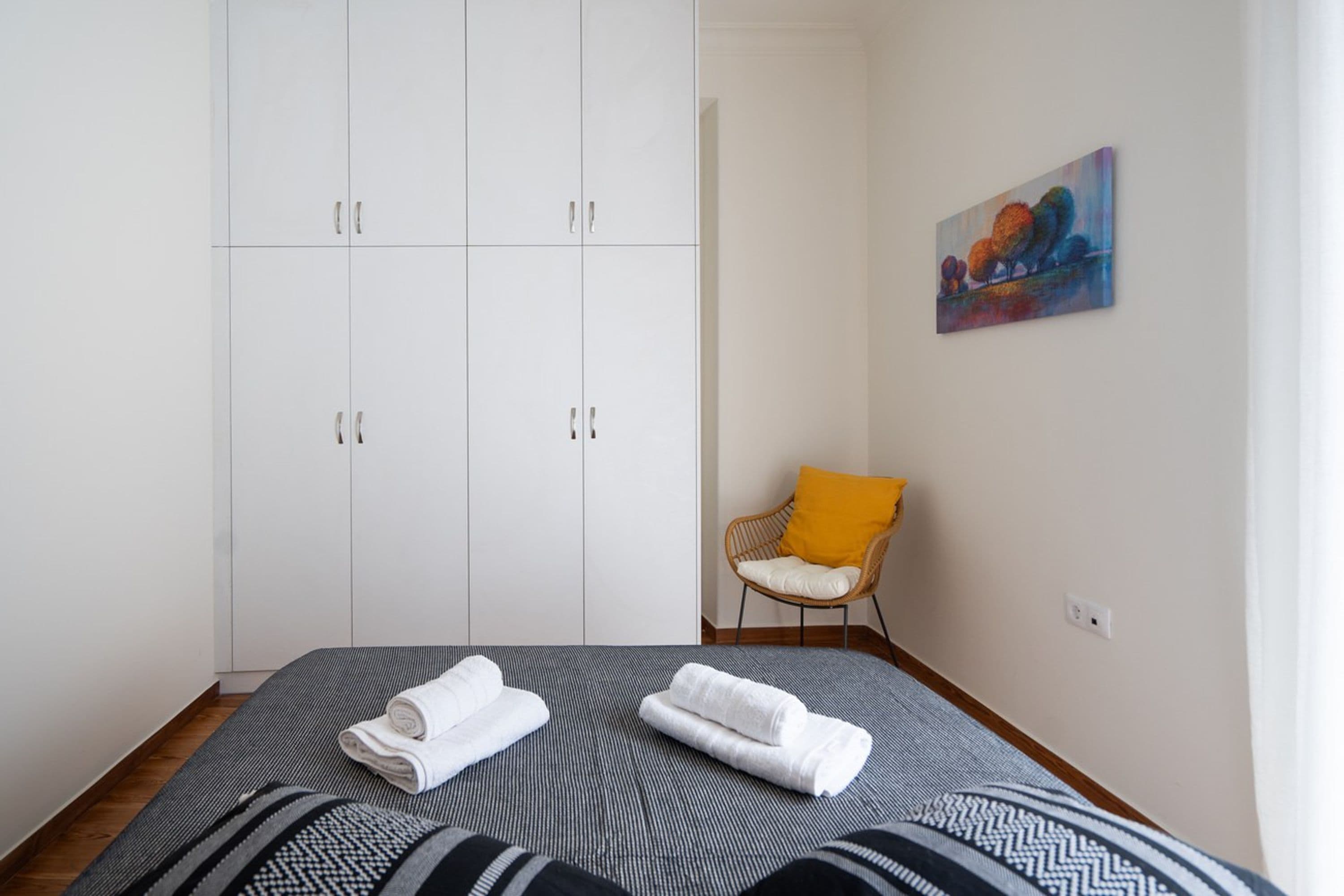 A bedroom with a bed, white wardrobe, a chair with a yellow cushion, and a painting on the wall.