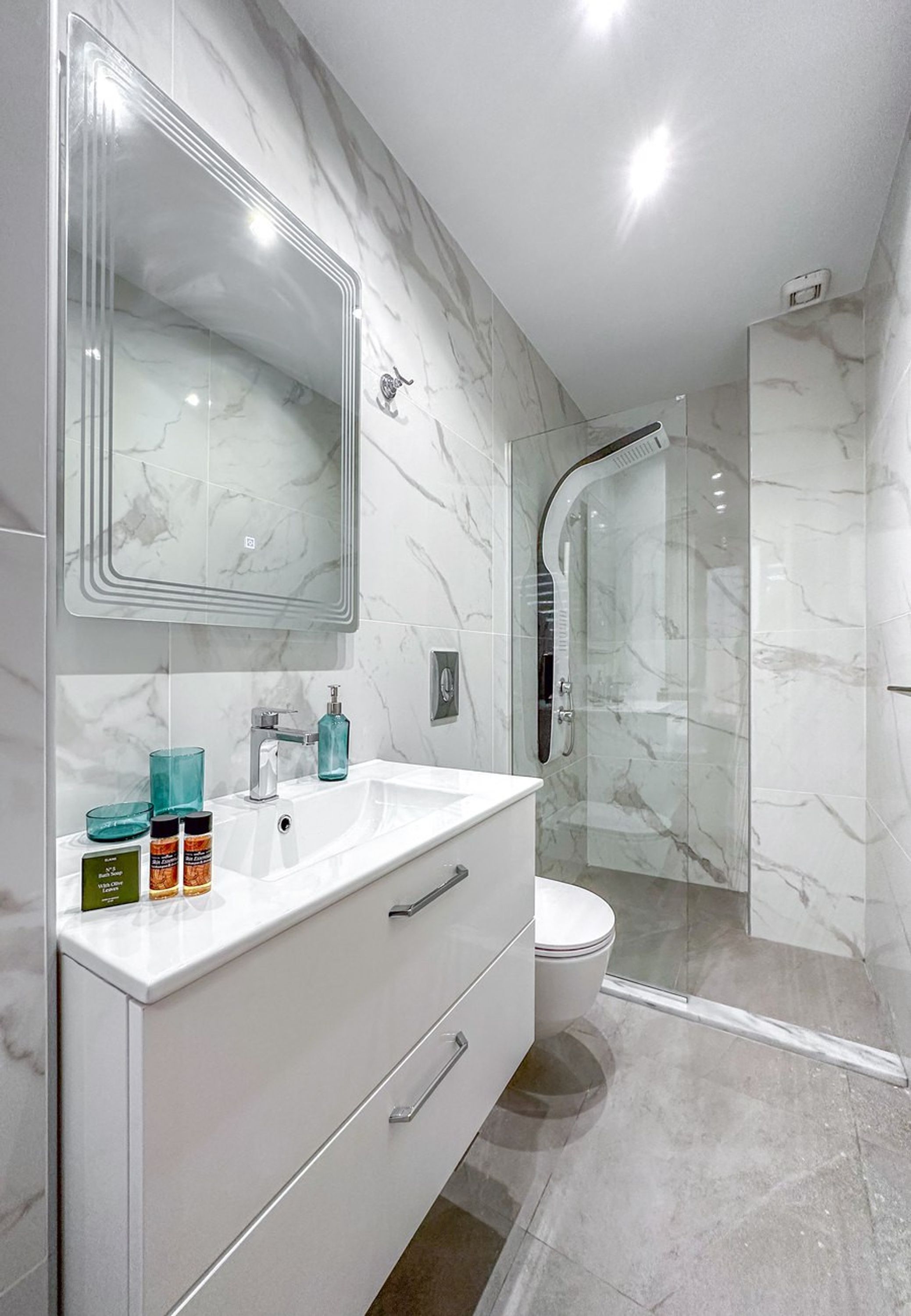 A modern bathroom with a white vanity, a large mirror, a glass shower enclosure, and marble walls.