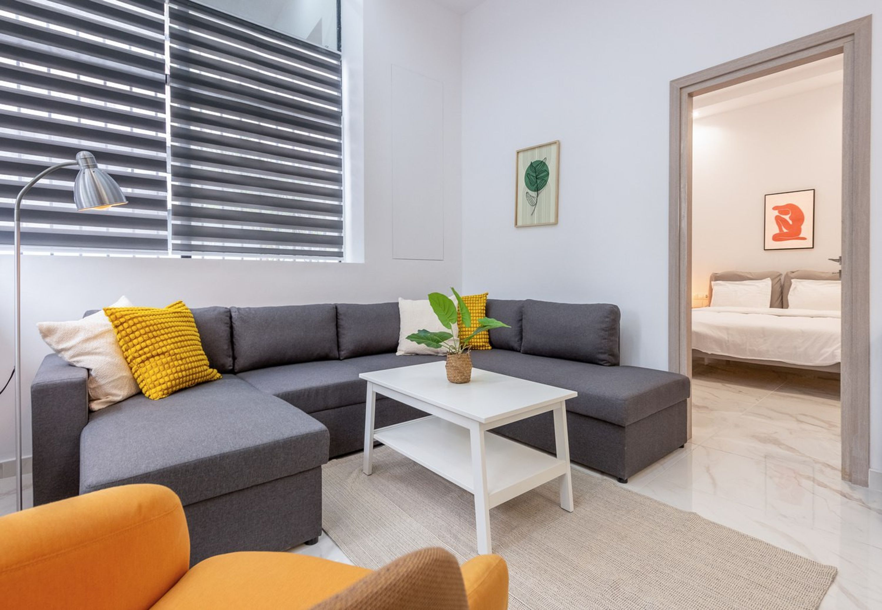 Una sala moderna con un sofá seccional gris, una mesa de centro blanca y una planta pequeña. Se ve una ventana con persianas, una lámpara y una cama al fondo.
