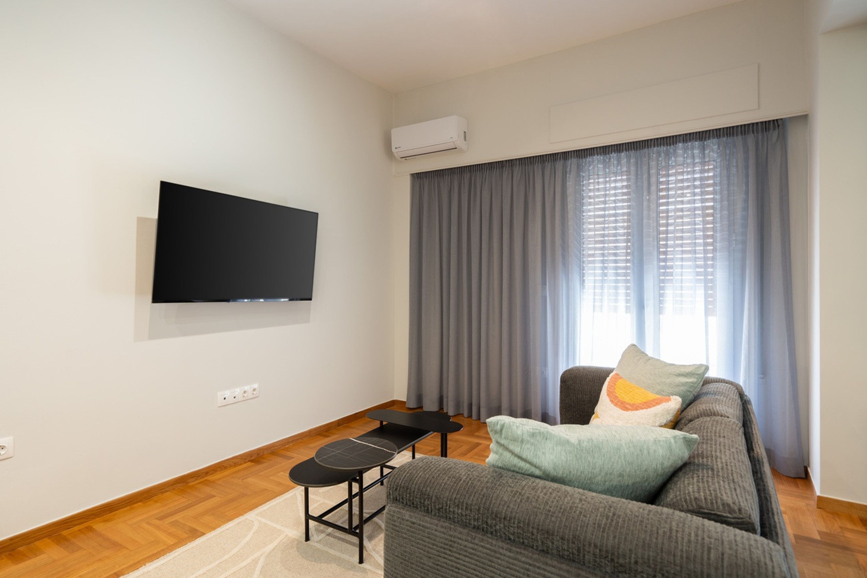 Ein Wohnzimmer mit Wandfernseher, einem grauen Sofa mit Kissen, einem kleinen schwarzen Tisch und einem Fenster mit grauen Vorhängen.