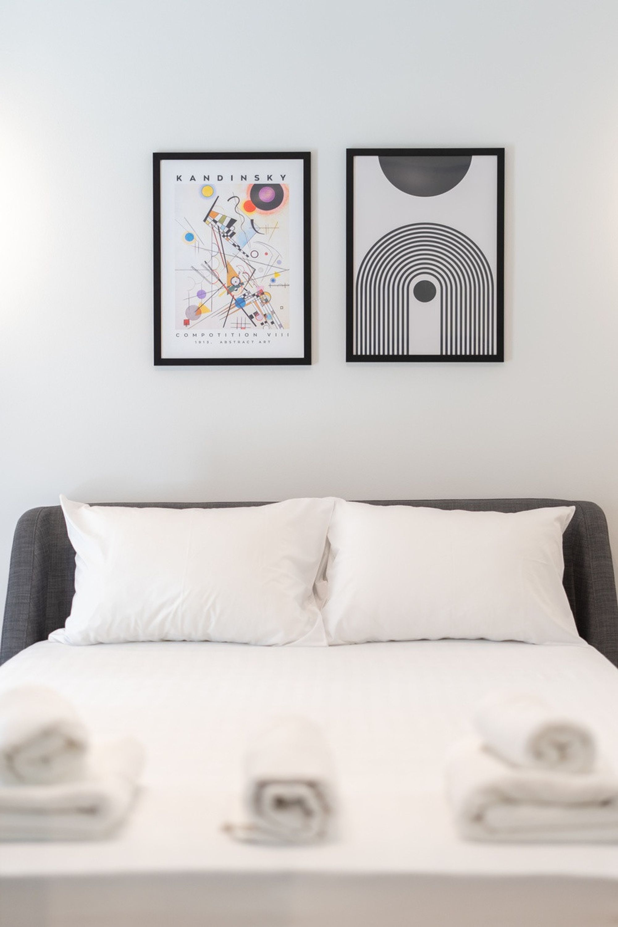 A neatly made bed with white linens and two pillows, a framed abstract art piece on the wall, and a framed geometric art piece.