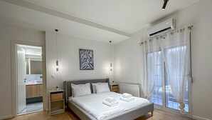 A modern bedroom with a large bed, bedside tables, a nightstand, a window with sheer curtains, and an air conditioning unit. - LIV - Modern Comfort Oasis in Pagrati (Kaisariani)
