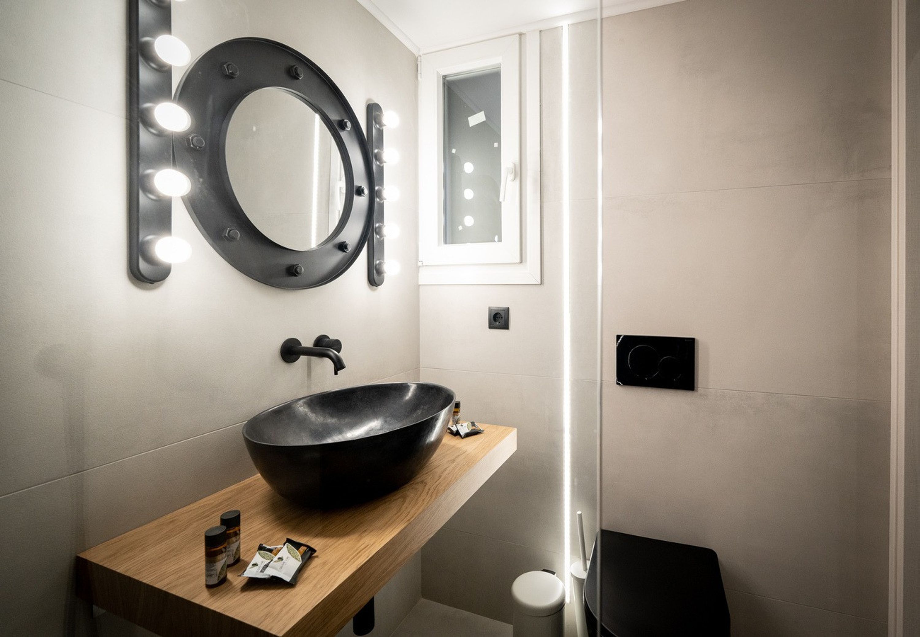Ein modernes Badezimmer mit einem großen, ovalen Waschbecken, einem runden Spiegel mit Beleuchtung und einem kleinen Fenster.