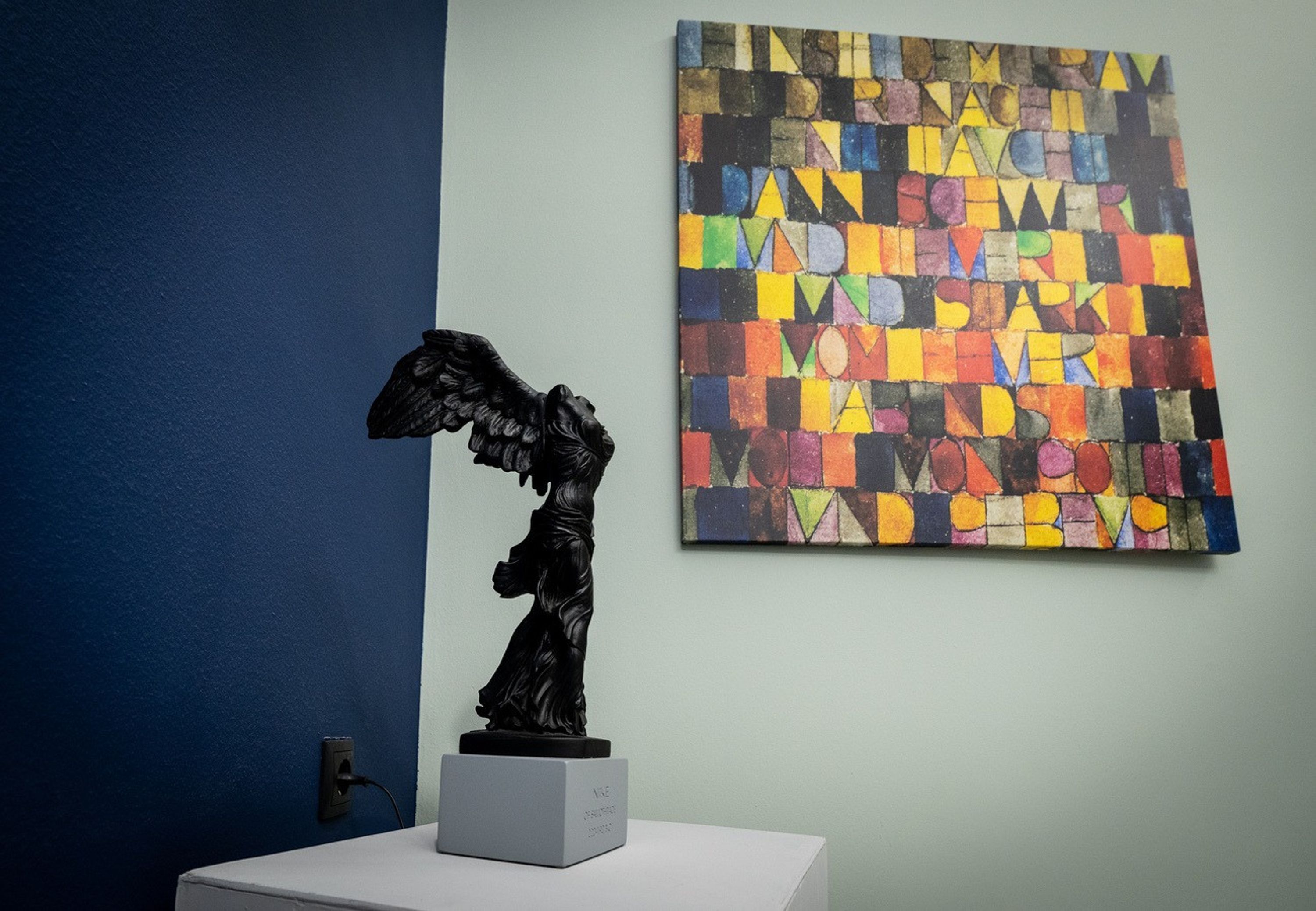 Una estatua sobre un pedestal en una habitación con pared azul y un cuadro abstracto colorido.