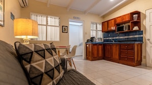 A living room with a sofa, a table, and a lamp. There is a kitchen area with wooden cabinets and a microwave. - Villas Paraiso B52 (Jacó)