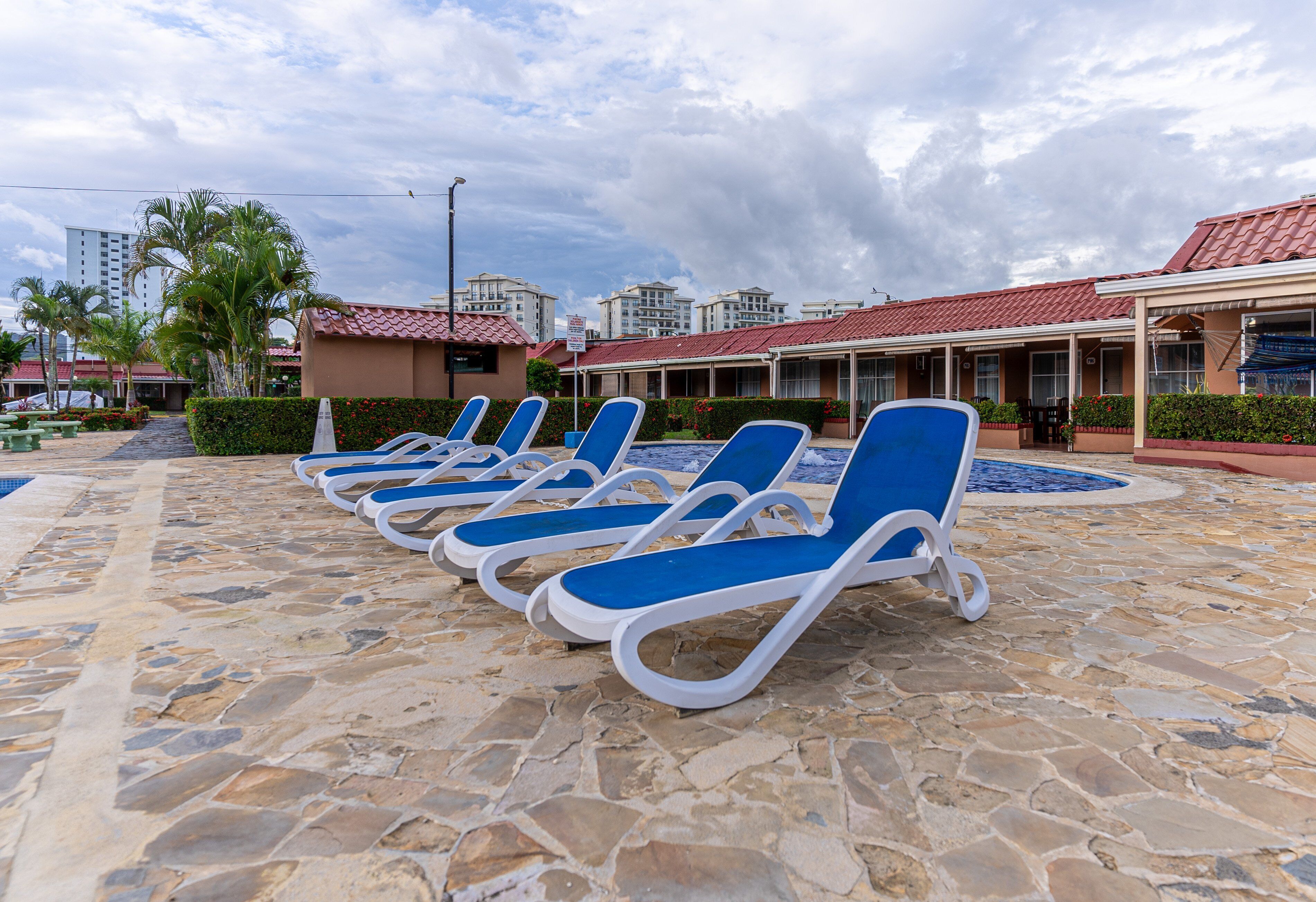 A poolside area with lounge chairs, a stone floor, and a building with a red-tiled roof in the background.