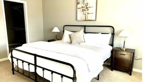A bedroom with a metal bed frame, white bedding, two bedside tables with lamps, a closet, and a wall-mounted abstract painting.
