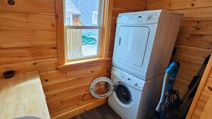 Een kleine wasruimte met houten wanden, een wasmachine, een droger en een raam met uitzicht op een huis.