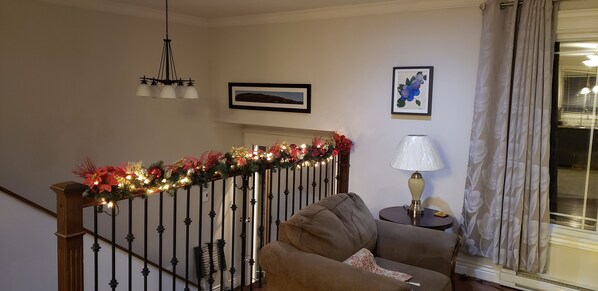 Una sala de estar con sofá, lámpara, escalera adornada con luces festivas y ventana con cortinas.