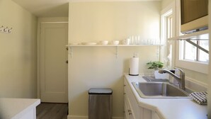 Cocina compacta con lavamanos, un cubo de basura, una ventana y una repisa con platos y tazas.