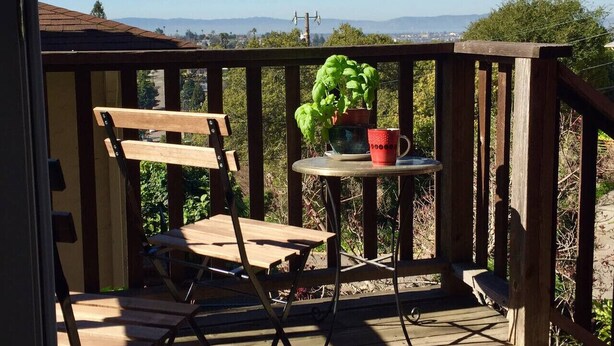 Una terraza de madera con una silla plegable y una mesita con una planta en maceta y una taza roja.