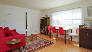Un espacio habitable compacto con un sofá rojo, una mesita redonda pequeña, una estantería, una mesa de comedor con sillas rojas y un amplio ventanal que proporciona luz natural.