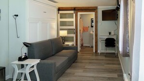 A modern living room with a gray sofa, a white side table, and a small white chair. A wooden door with frosted glass panels leads to a bathroom with a towel hanging on the wall. - Beach Bum Hideaway - Cozy studio condo with full kitchen. 400 ft to the beach! (Hampton)