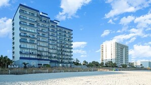 Un hotel frente a la playa con varios pisos, balcones y una playa arenosa al frente.