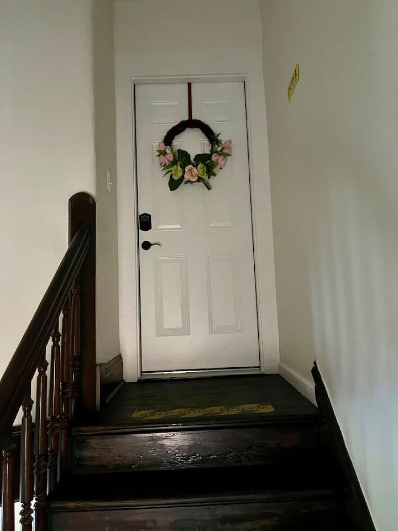 A white door with a floral wreath, a black doorbell, and a wooden staircase with a dark finish.