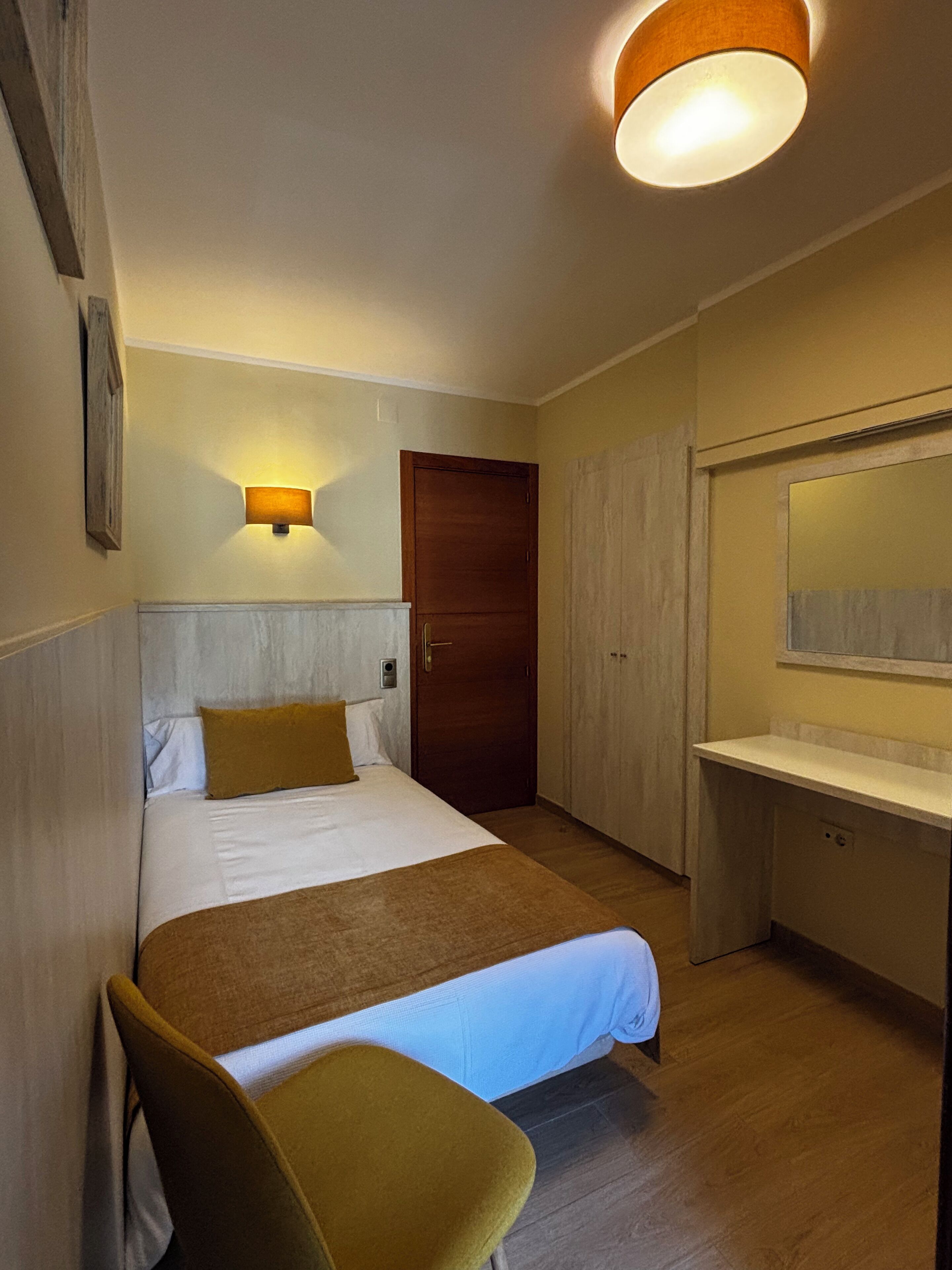 Una habitación de hotel con una cama, una mesita de noche, una silla, un armario y un espejo.