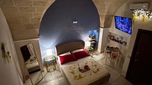 Une chambre avec un mur en pierre, un lit avec des coussins rouges, un téléviseur fixé au mur et une petite table avec une lampe.