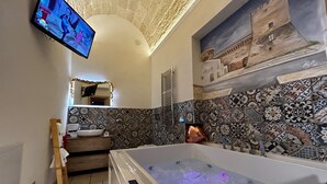 Une salle de bain dotée d’une grande baignoire, d’un téléviseur fixé au mur et d’un mur recouvert de carreaux orné d’une fresque représentant un château.