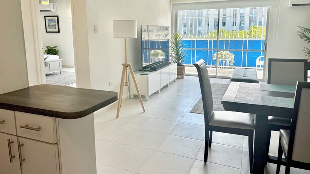 A modern living room with a dining area, white walls, and a large window offering a view of the outside. - Modern 1 BR Condo at ISLA VERDE, Free parking, Pool & Wifi (Carolina)