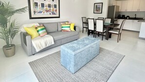 A modern living room with a grey sofa, colorful cushions, a blue ottoman, and a dining area with wooden chairs and a table. - Modern 1 BR Condo at ISLA VERDE, Free parking, Pool & Wifi (Carolina)
