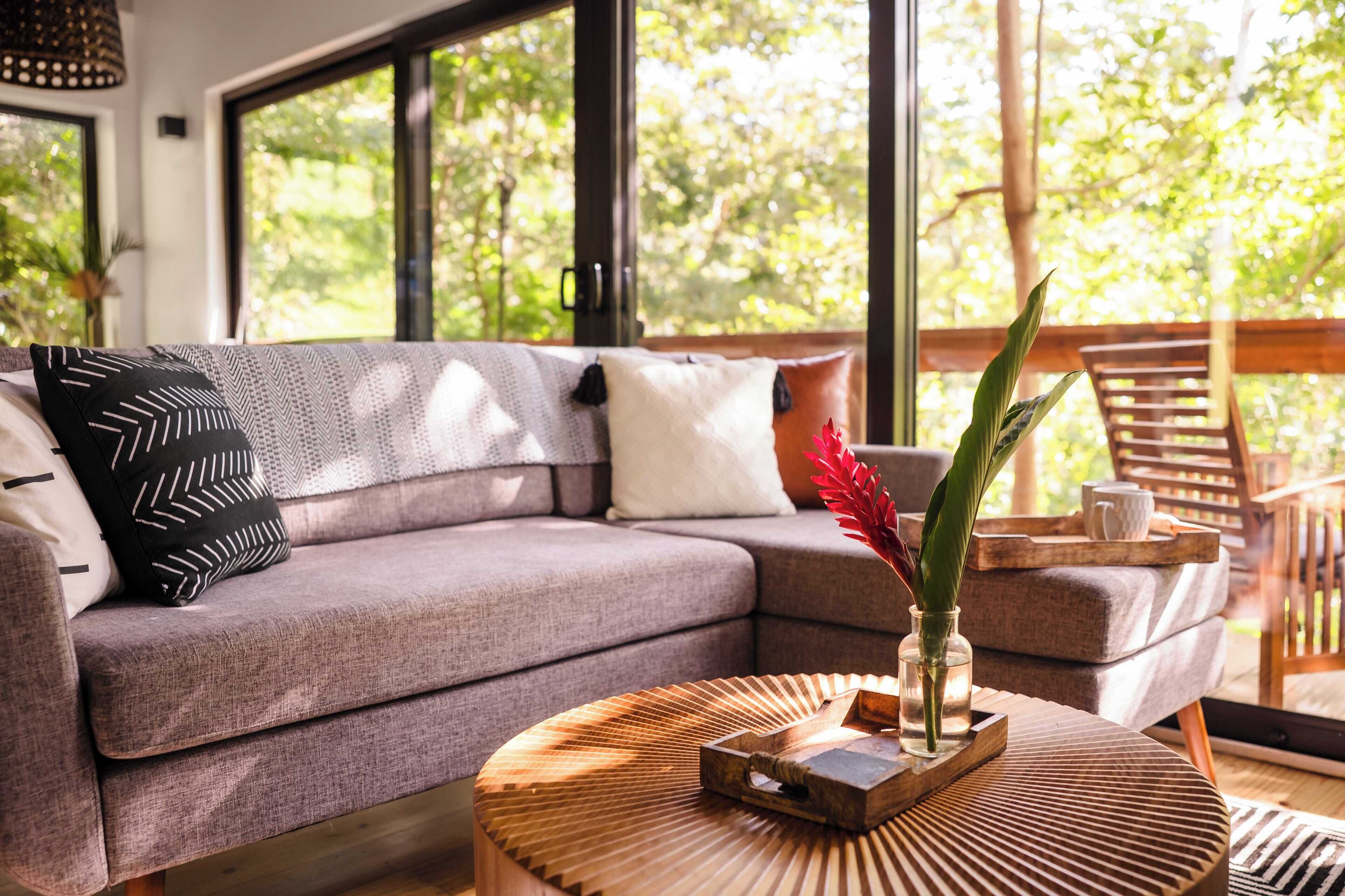 A modern living room with a grey sofa, a round wooden coffee table, and a view of greenery through large windows.