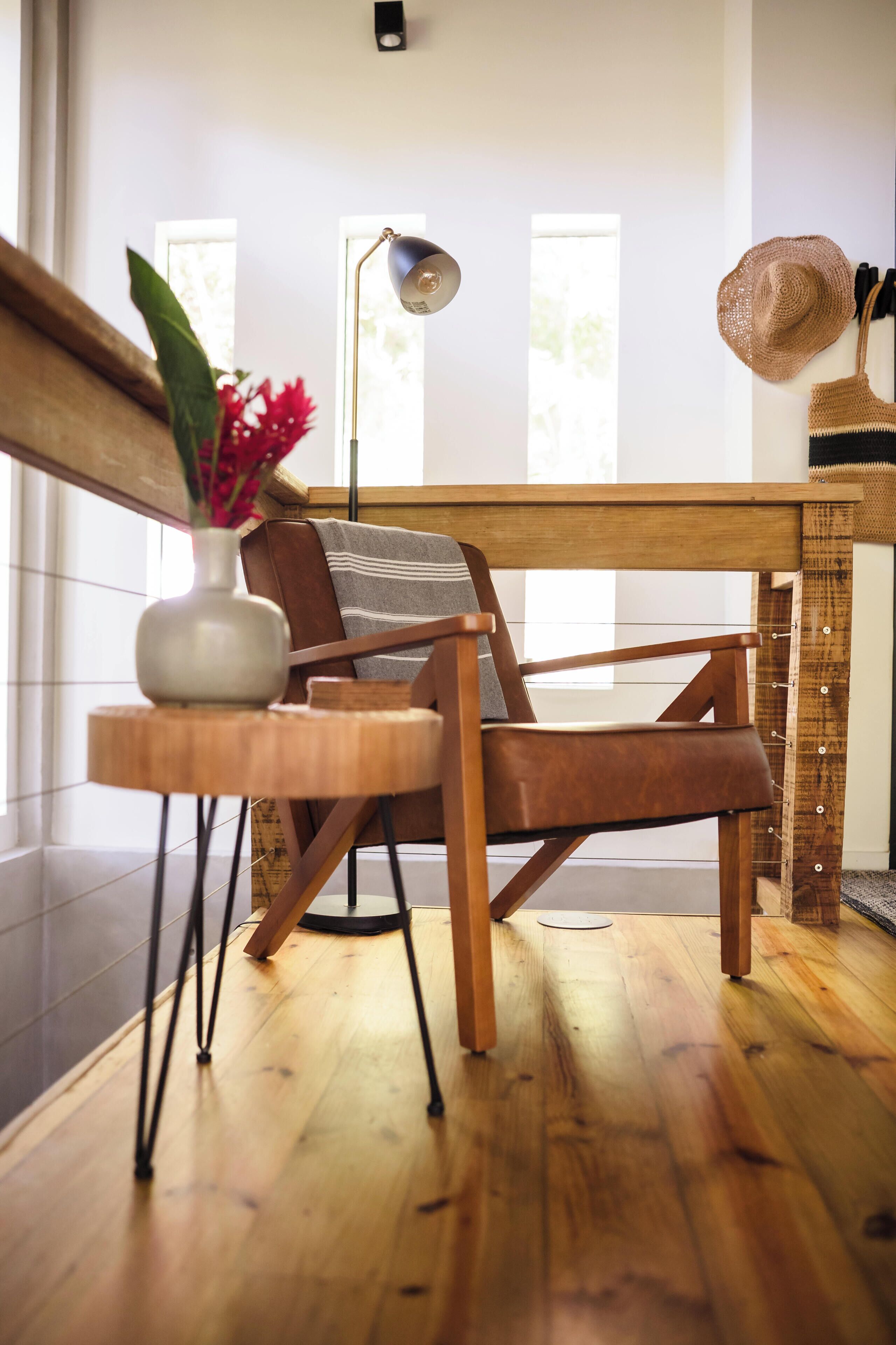 Een houten stoel met kussen, een klein rond houten tafeltje met een bloemenvaas, en een vloerlamp in een kamer met houten vloer en een raam.