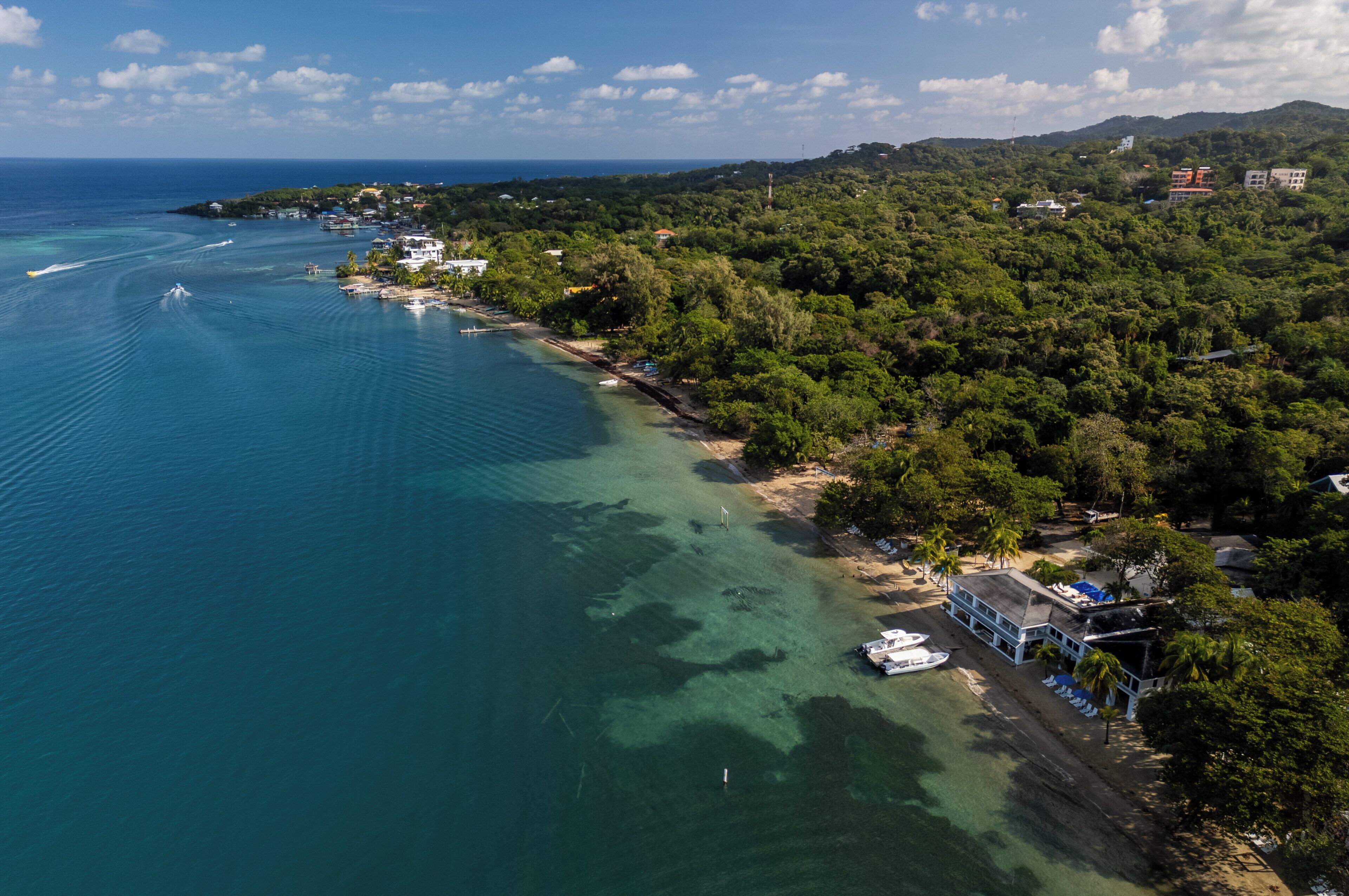 An aerial view of a coastal area with clear waters, a beach, and a mix of residential and commercial buildings.